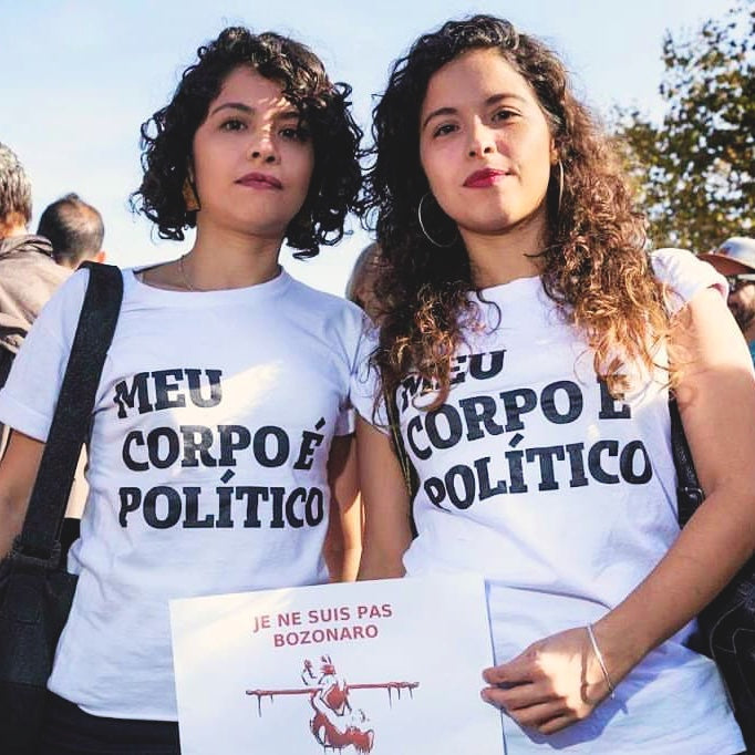 Foto colorida de duas mulheres brancas de cabelos cacheados, a da esquerda curto e da direita longo, ambas vestem camiseta branca escrito meu corpo é político em preto,, fundo céu e folhagem