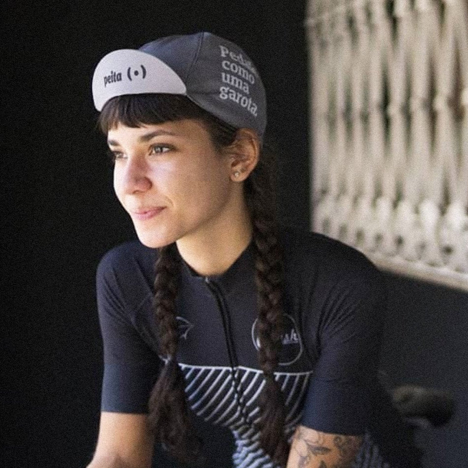 Foto colorida de uma mulher branca, cabelos castanhos, longos, com 2 tranças, com cap cinza escrito pedale como uma garota em branco do lado direito, e a logo e palavra peita escrito em preto. Está na bike, olhando para frente.