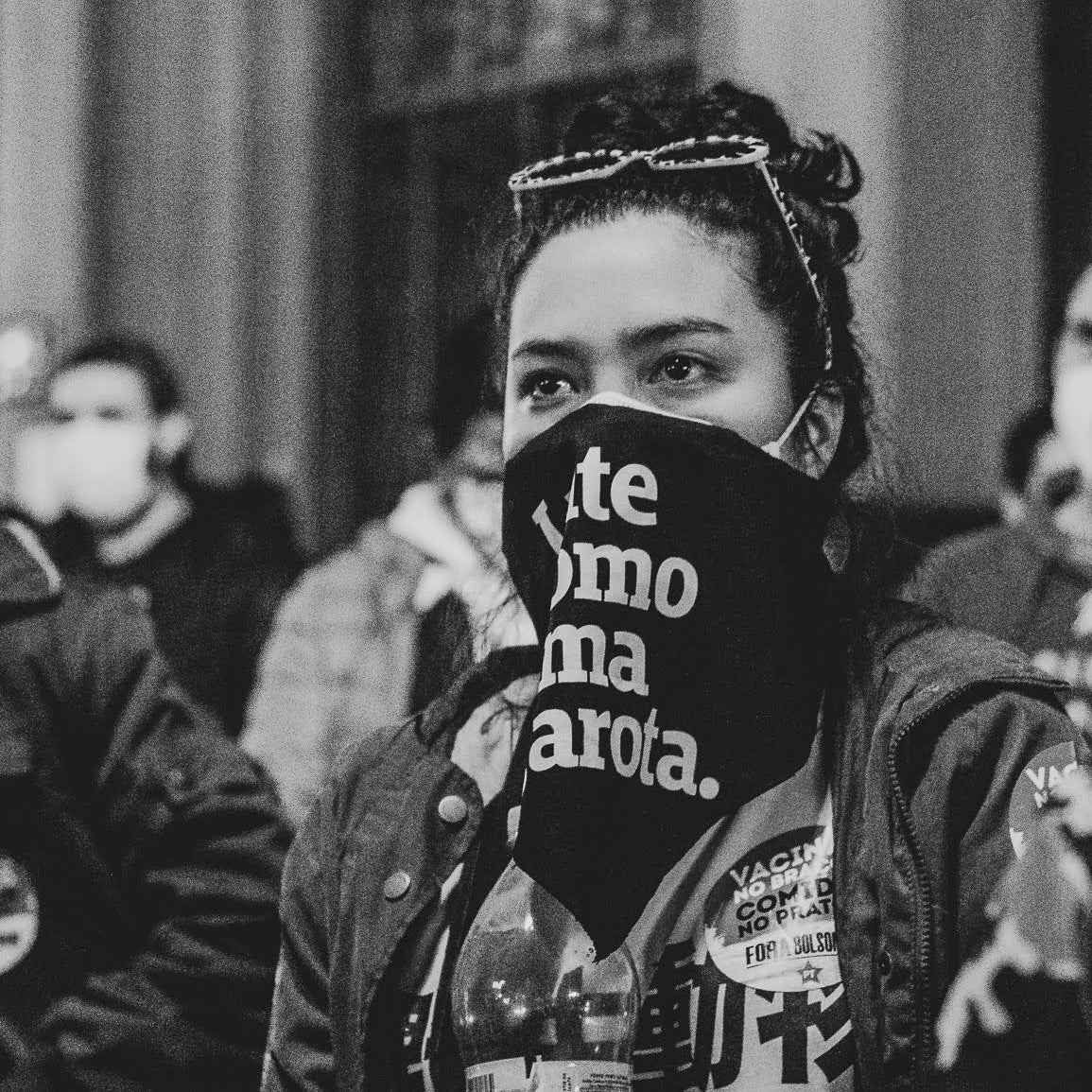 Foto preto e branco de uma mulher branca com cabelos escuros presos, com um lenço preto escrito lute como uma garota em branco. Está em uma manifestação.