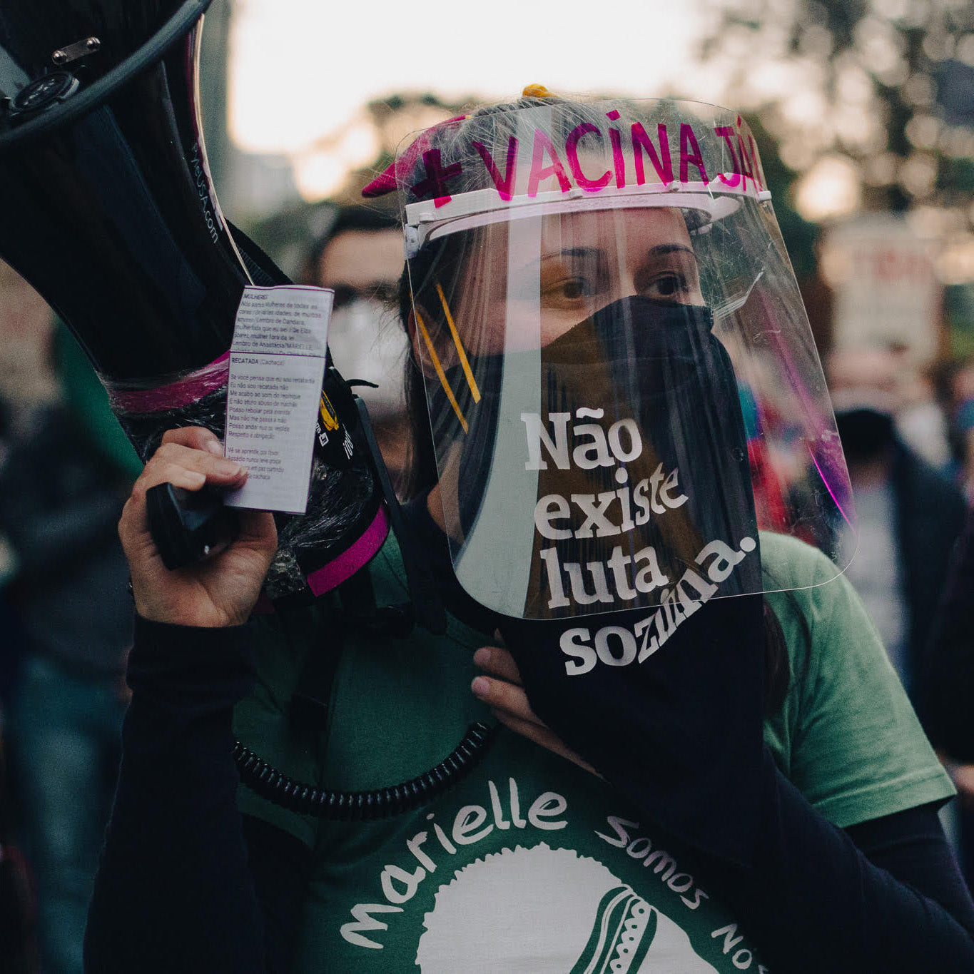 Foto colorida de uma mulher branca de cabelos castanhos e presos, usa um lenço de guerrilha preto escrito Não existe luta sozinha em branco, está com uma  máscara de proteção facial transparente e segura um megafone