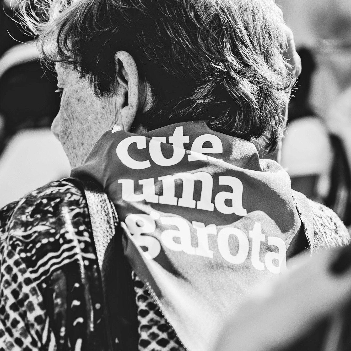 Foto preta e branca de uma senhora branca de cabelos grisalhos, curtos e tem um lenço cinza amarrado no pescoço, escrito lute como uma garota em branco. Ela usa uma bolsa no ombro esquerdo, está de costas para a foto.