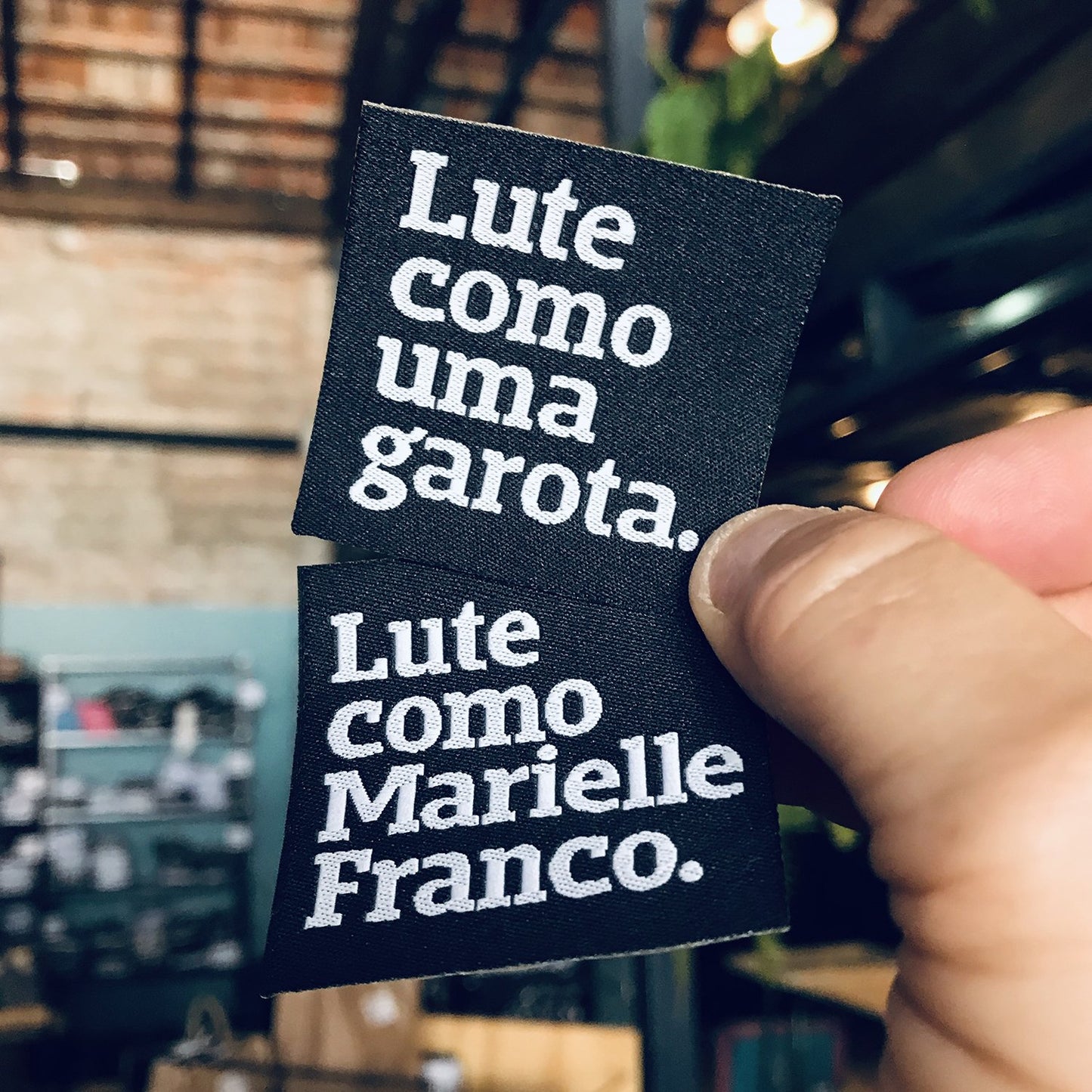 Foto preta e branca de patch pretocom frase seja quem você quiser em branco. Foto colorida de uma mão branca segurando 2 patches pretos com as frases lute como uma garota e lute como Marielle Franco em branco.