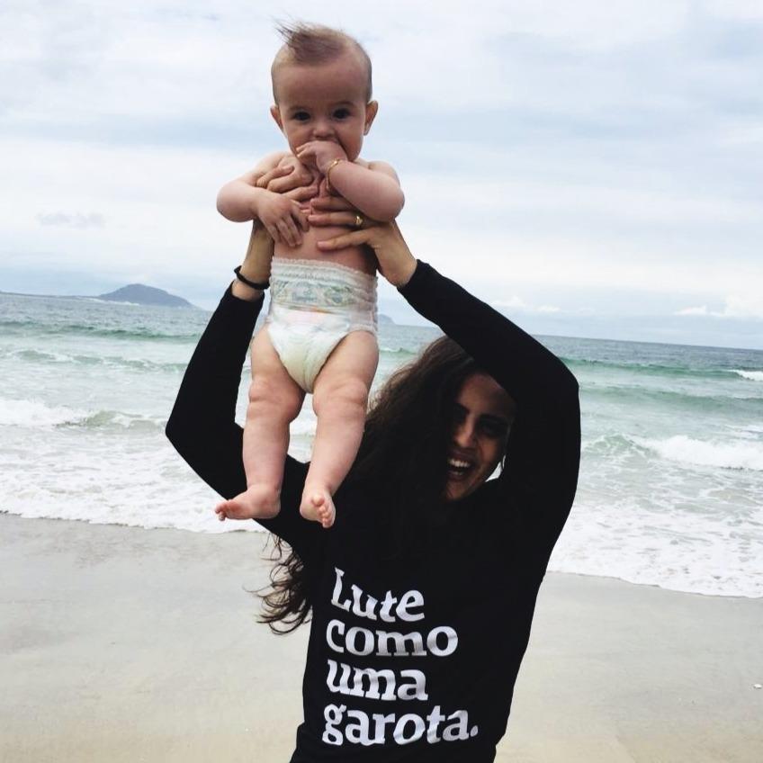 Foto colorida de uma mulher branca, com cabelos castanhos, longos, soltos, ondulados, vestida com rash guard de manga comprida, preta, escrita lute como uma garota em branco. Ela segura em suas mãos uma criança branca com fralda descartável sorri, está na praia.