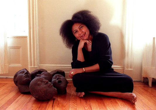 Foto mulher negra sentada em uma sala com cabeças de estátuas marrom. bell é negra, com cabelos crespos médios está vestida de preto.  