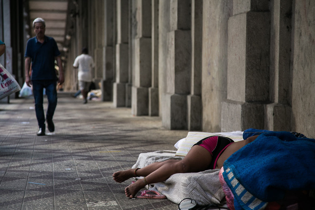 foto colorida em ambiente externo de uma mulher negra deitada em um colchão na calçada. ela está deitada, coberta na cabeça e só se vê suas pernas e quadril. usa um short preto. 