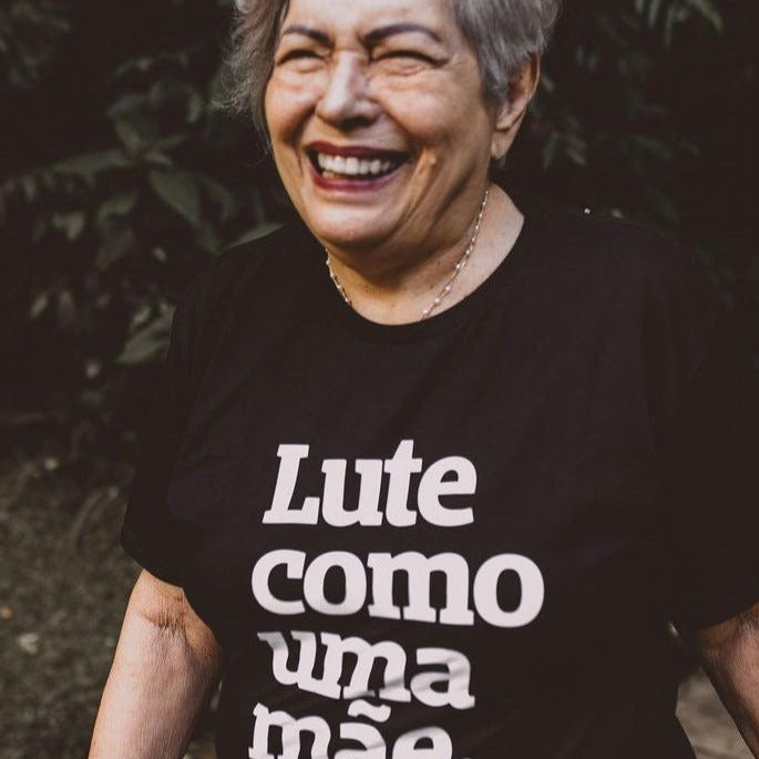 foto colorida e externa. Dona Lourdes está no parque e veste camiseta preta da peita com a frase "lute como uma mãe" estampada em branco. ela tem cabelo curto e grisalho e está sorrindo para a foto.