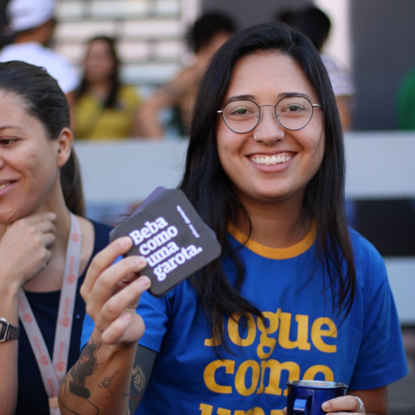 Foto colorida de uma mulher branca de cabelo castanho, liso, com franja comprida e veste  uma camiseta azul escrito jogue como uma garota amarela. Ela segura  um porta copo preto escrito Beba como uma garota em branco, posicionado em frente ao seu olho esquerdo. Tem tatuagens no braço e está sorrindo.