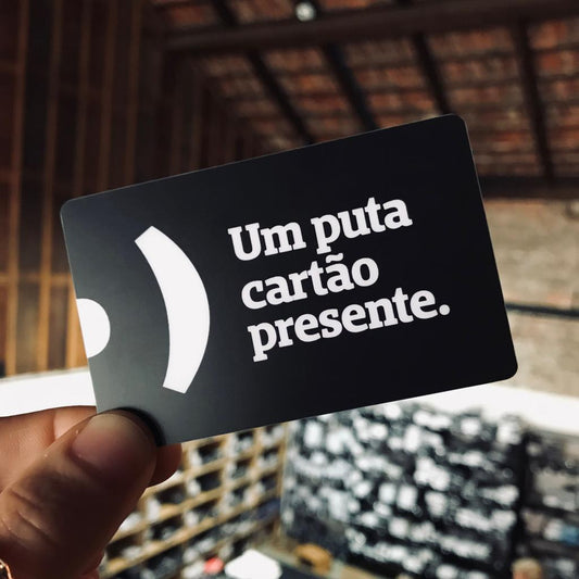Foto colorida de um cartão preto com a logo da Peita, com a frase  um puta cartão presente em branco. Está sendo segurado por uma mão branca.