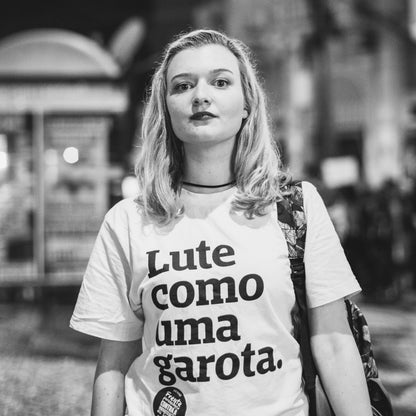 Foto preto e branco de uma mulher branca de cabelos curtos e loiros, veste camiseta branca com a frase Lute como uma garota em preto, com mochila pendurada no braço direito. Fundo desfocado