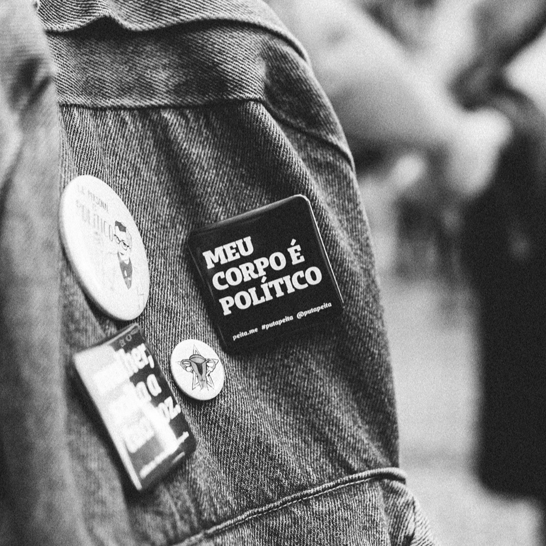 Foto preta e branca de uma mochila jeans com um  botton preto, quadrado, com  a frase meu corpo é político em branco. 