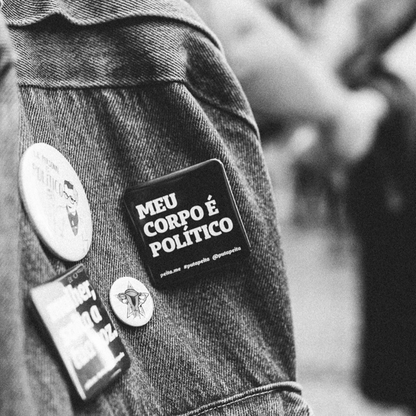 Foto preta e branca de uma mochila jeans com um  botton preto, quadrado, com  a frase meu corpo é político em branco. 