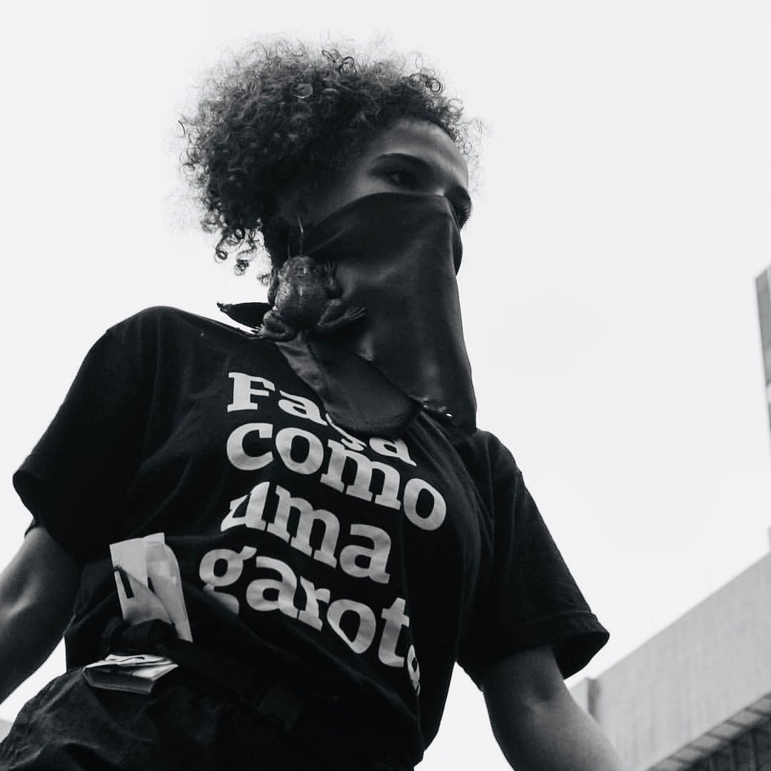 Foto branca e preta de uma mulher negra de cabelos crespos, escuros, presos em um rabo e veste uma camiseta preta escrita faça como uma garota em branco. Ela tem uma bandana cobrindo a boca e na sua orelha usa um brinco em formato de sapo. Olha pra frente, séria.