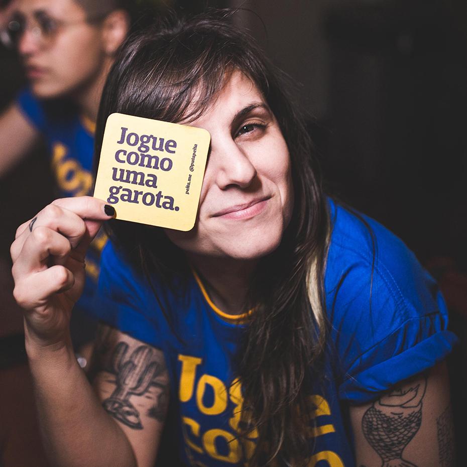 Foto colorida de uma mulher branca de cabelo castanho, liso, com franja e veste  uma camiseta azul escrito jogue como uma garota em amarelo. Ela segura  um porta copo amarelo escrito jogue como uma garota em azul, posicionado em frente ao seu olho esquerdo. Tem  tatuagens no braço e está sorrindo.