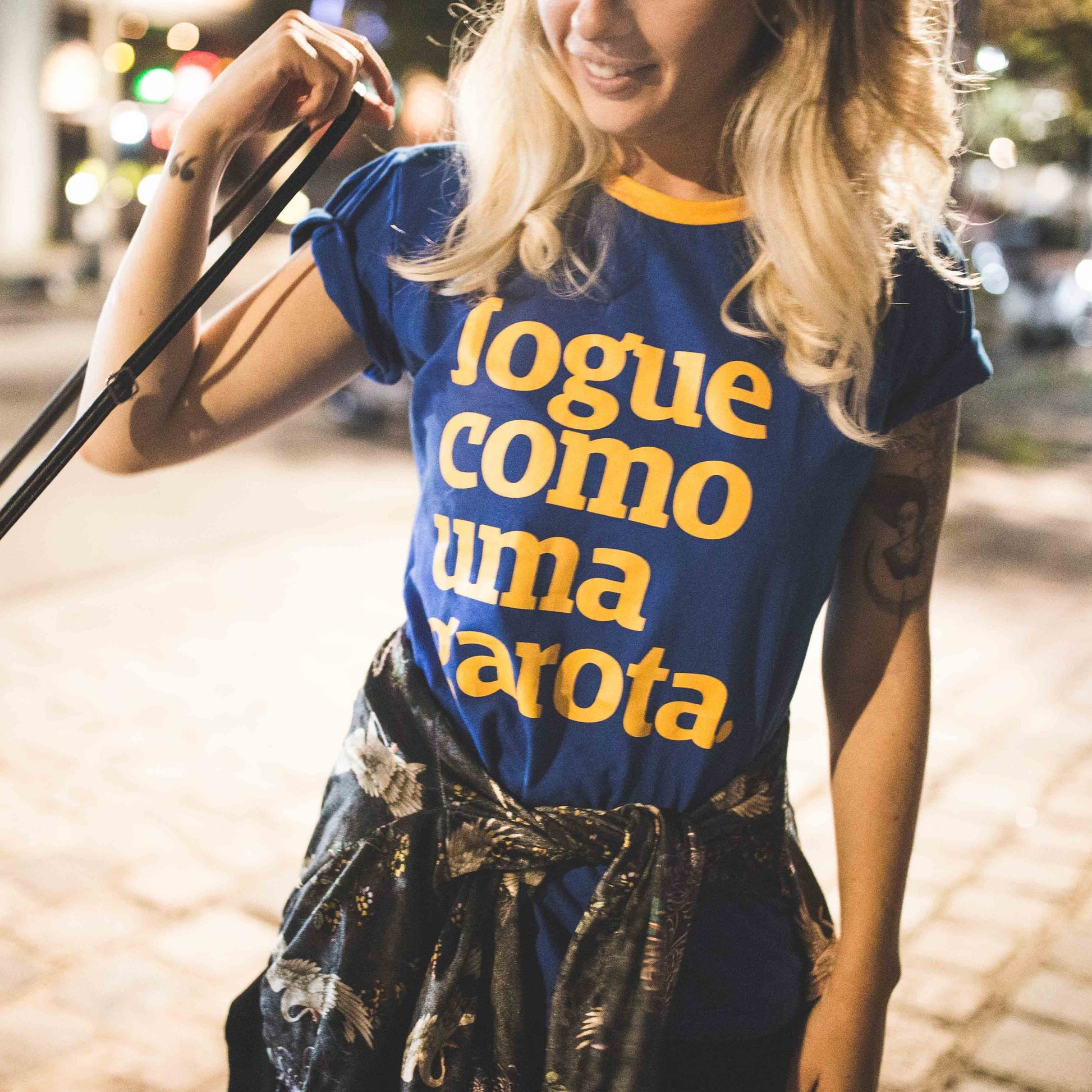 Foto colorida de uma mulher branca de cabelos loiros, soltos,lisos e compridos. Está vestindo uma camisa azul escrito jogue como uma garota amarela. Ela tem uma blusa amarrada na cintura e está com sua bolsa no ombro esquerdo, sorri na foto.