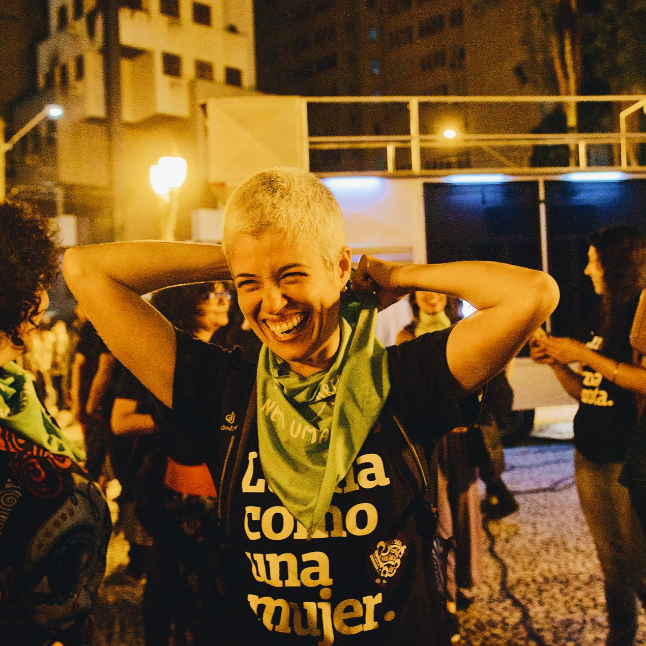 Foto colorida de uma mulher branca, cabelo raspado, loiro, com camiseta preta escrita lucha como una mujer em branco, com mochila nas costas, descontraída, sorrindo na foto.