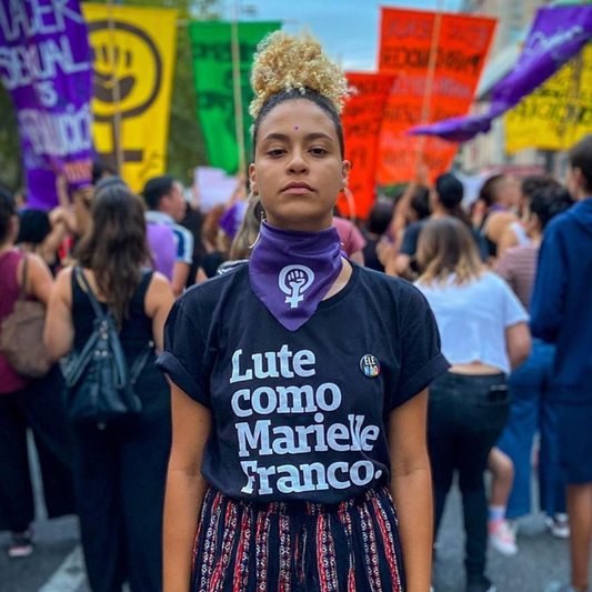 Foto colorida de uma mulher negra, cabelos crespos, castanhos com pontas loiras, vestida com camiseta preta escrito lute como Marielle Franco em branco. Ela está parada em uma multidão e olha séria para foto.