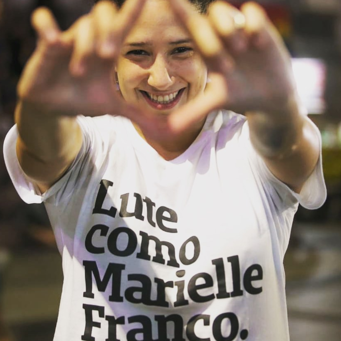 Foto colorida de uma mulher branca, cabelos castanhos, cacheados, estão presos para trás, vestida com camiseta branca escrito lute como Marielle Franco em preto.As mãos fazem gesto imitando uma vulva, sorri na foto.