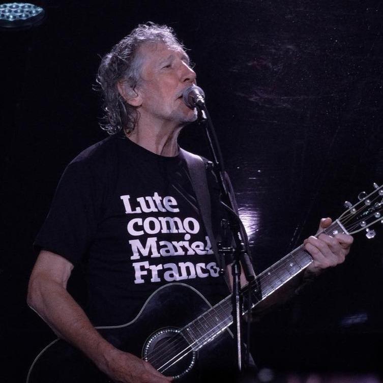 Foto colorida  do Roger Waters, homem branco, com cabelos , grisalhos, com camiseta preta escrito lute como Marielle Franco em branco, toca  um violão, enquanto canta no microfone.