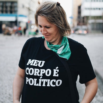 Foto colorida de uma mulher branca, cabelos loiros, lisos, chanel, está vestida com camiseta preta escrito meu corpo é político em branco. Ela olha pra chão com leve sorriso.