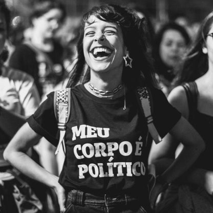 Foto preta e branca de uma mulher branca, cabelos castanhos, longos, com franja, lisos, meio soltos, usa brinco estrelas na orelha esquerda, está vestida com camiseta preta escrita meu corpo é político em branco. Ela sorri com as mãos na cintura. 