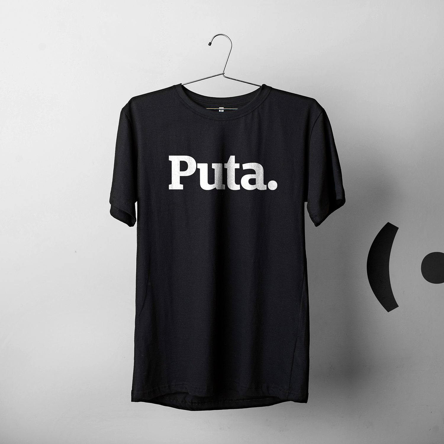 Foto colorida de uma camiseta preta escrito Puta em branco, fundo cinza com logo da Peita.