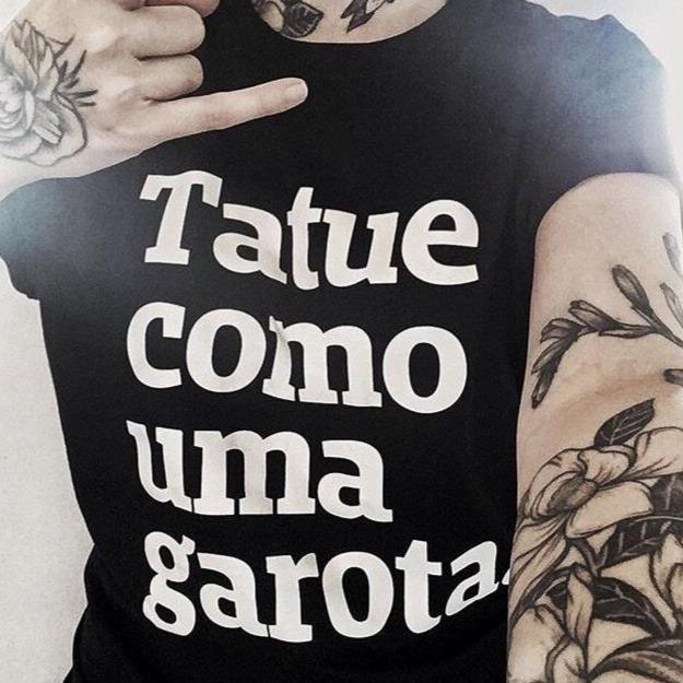 Foto selfie colorida do tronco de uma mulher branca, vestida com camiseta preta escrita tatue como uma garota em branco. Ela tem tatuagens nos braços e faz hang loose com a mão esquerda.