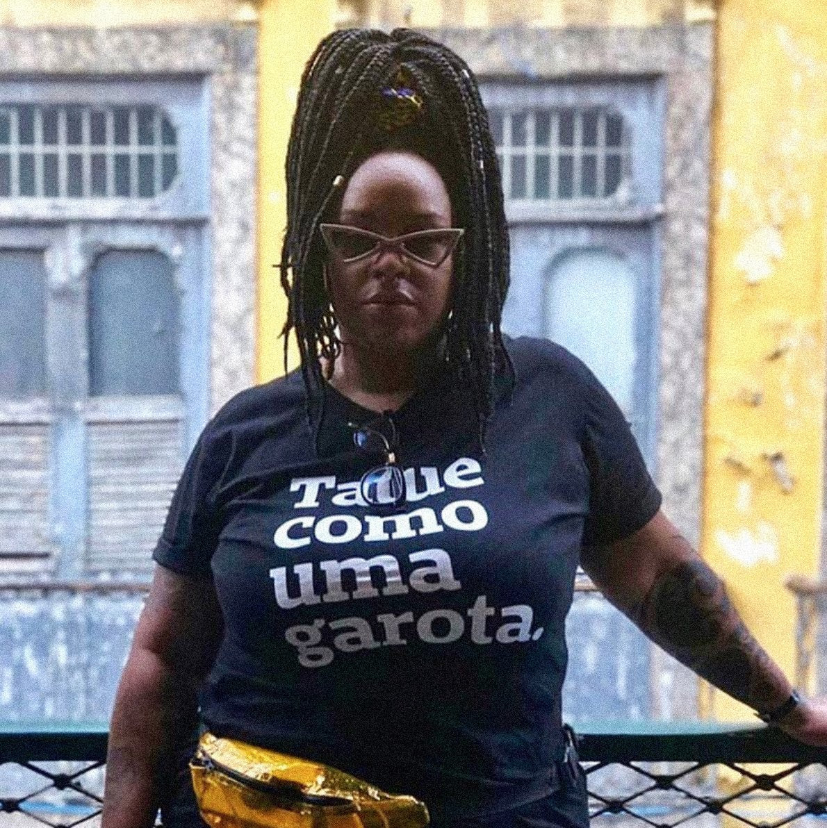 Foto colorida de uma mulher negra, com cabelos trançados, pretos, presos com rabo, usa óculos escuros, tem tatuagens nos braços, está vestida com camiseta preta escrita tatue como uma garota em branco e pochete amarela na cintura. Está encostada em uma grade de sacada