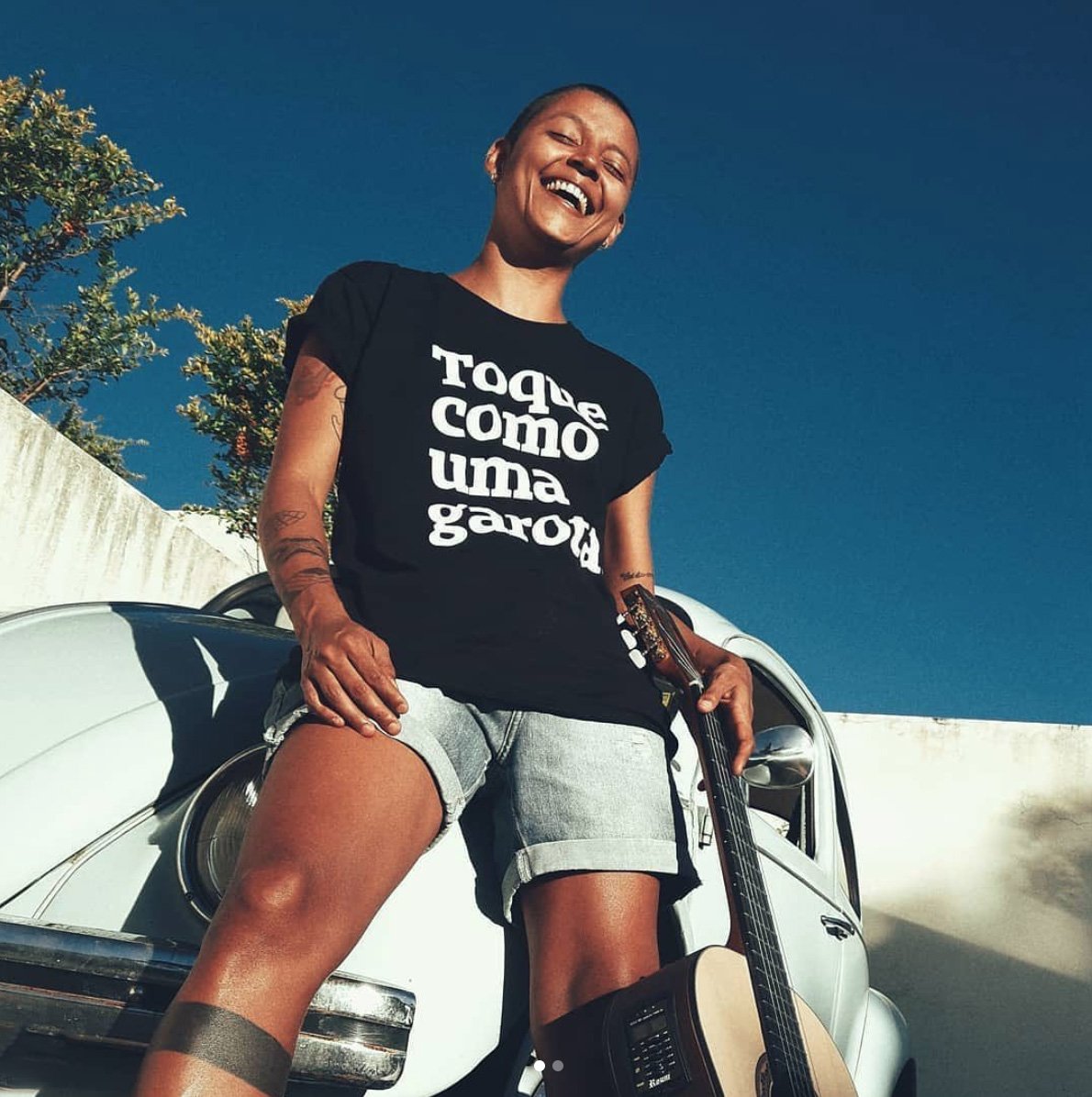 Foto colorida de uma mulher branca, cabelo escuro, raspado, com tatuagem no braço e perna, vestida com camiseta preta escrito toque como uma garota em branco. Na sua mão esquerda segura um violão na perna e está encostada em um fusca azul claro, sorri na foto.