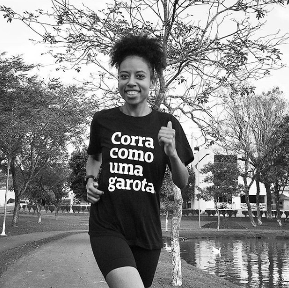 Foto preta e branca de uma mulher negra, com cabelos crespos, presos,  vestindo uma camiseta preta com a frase corra como uma garota em branco, short preto, correndo no parque com sorriso no rosto.