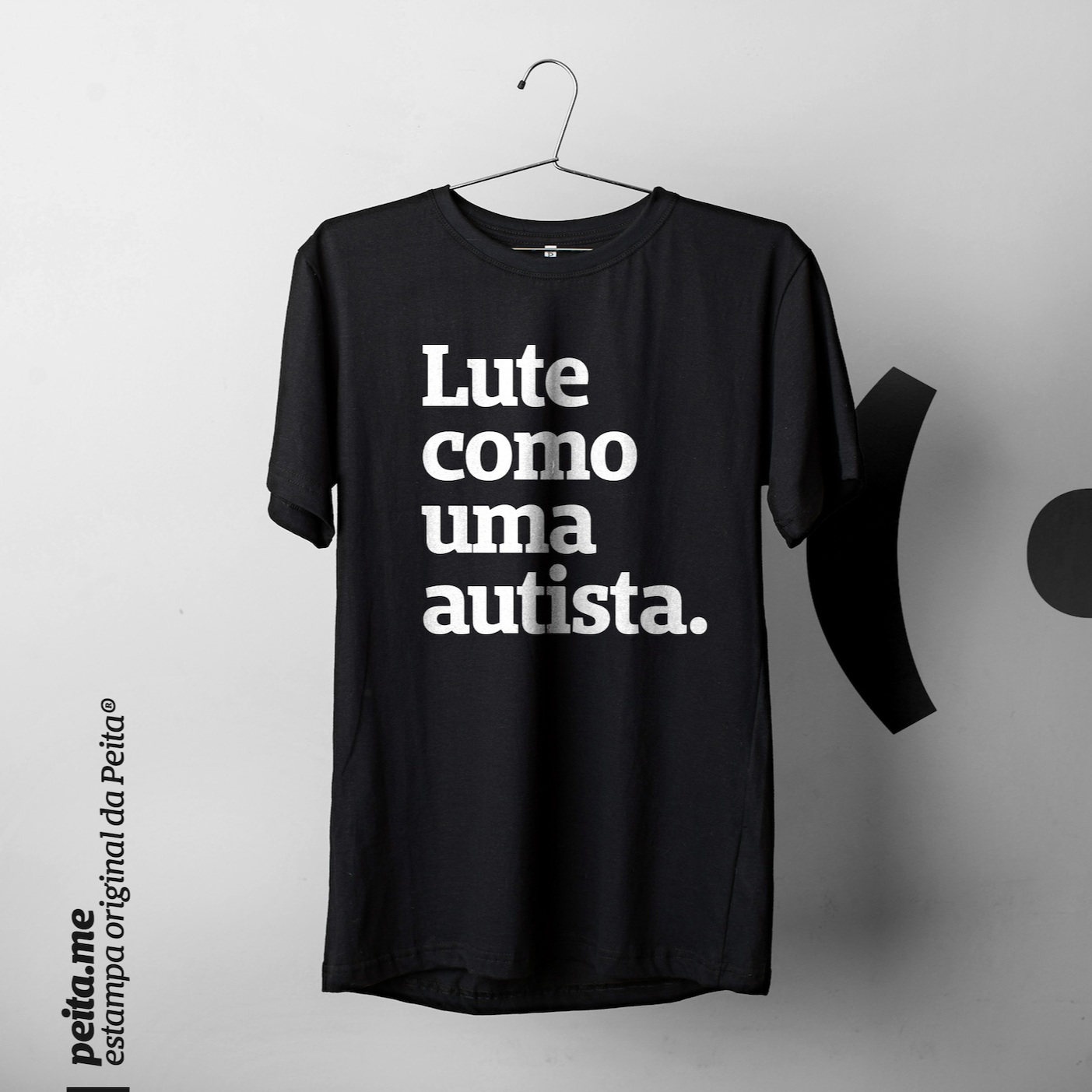 Foto preta e branca de uma camiseta preta com a frase lute como uma autista em estampada branco, pendurada em um cabide. Fundo cinza claro.
