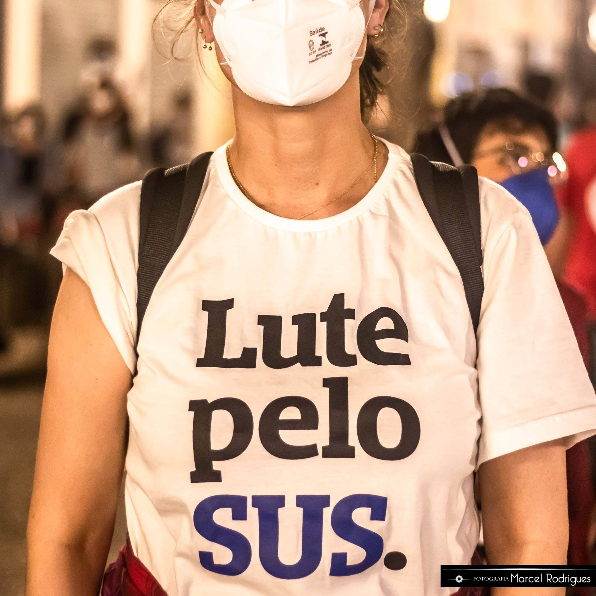 Foto colorida de mulher branca de cabelo liso preso, Veste uma camiseta branca escrito lute pelo sus em preto e azul, com mascara branca e mochila preta nas costas. Fundo desfocado