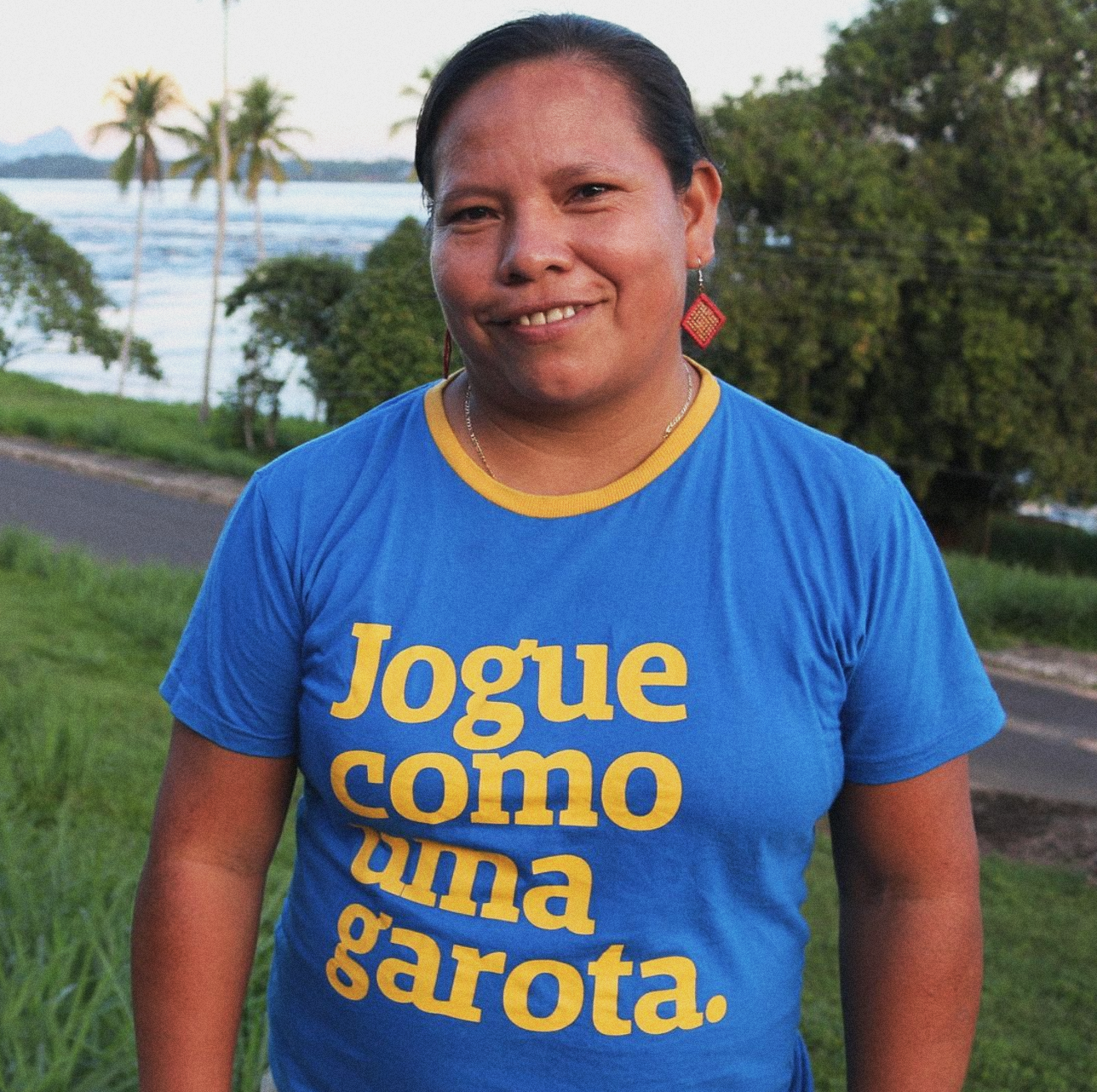 Foto colorida de mulher indígena de cabelos escuros que veste camiseta azul escrito Jogue como uma garota em amarelo com gola amarela. Usa brinco vermelho com detalhe em amarelo de forma geométrica. Ao fundo praia e natureza