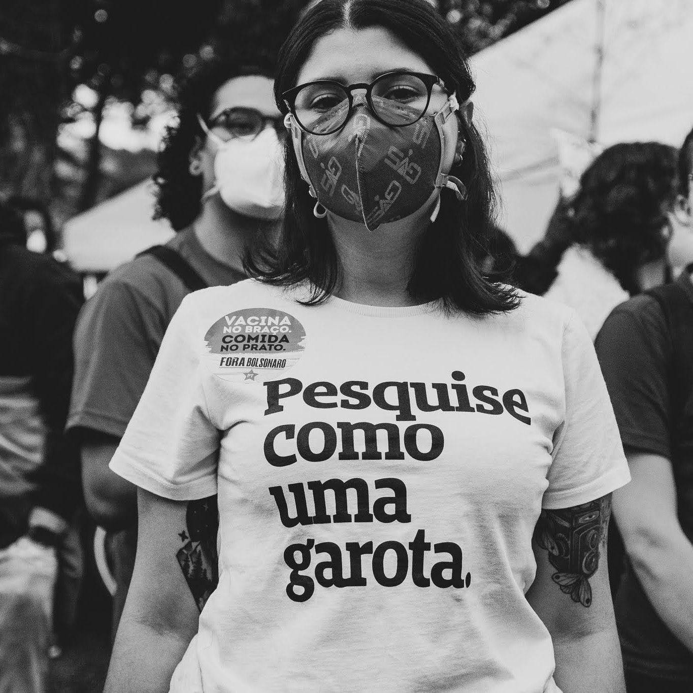 Foto preto e branco de mulher branca de cabelo escuro curto, veste uma camiseta branca com frase Pesquise como uma garota em preto, ela usa óculos arredondados, e um adesivo  com a frase Vacina no braço, comida no prato no canto superior esquerdo do peito, com tatuagens nos dois braços e mascara preta. Fundo uma manifestação