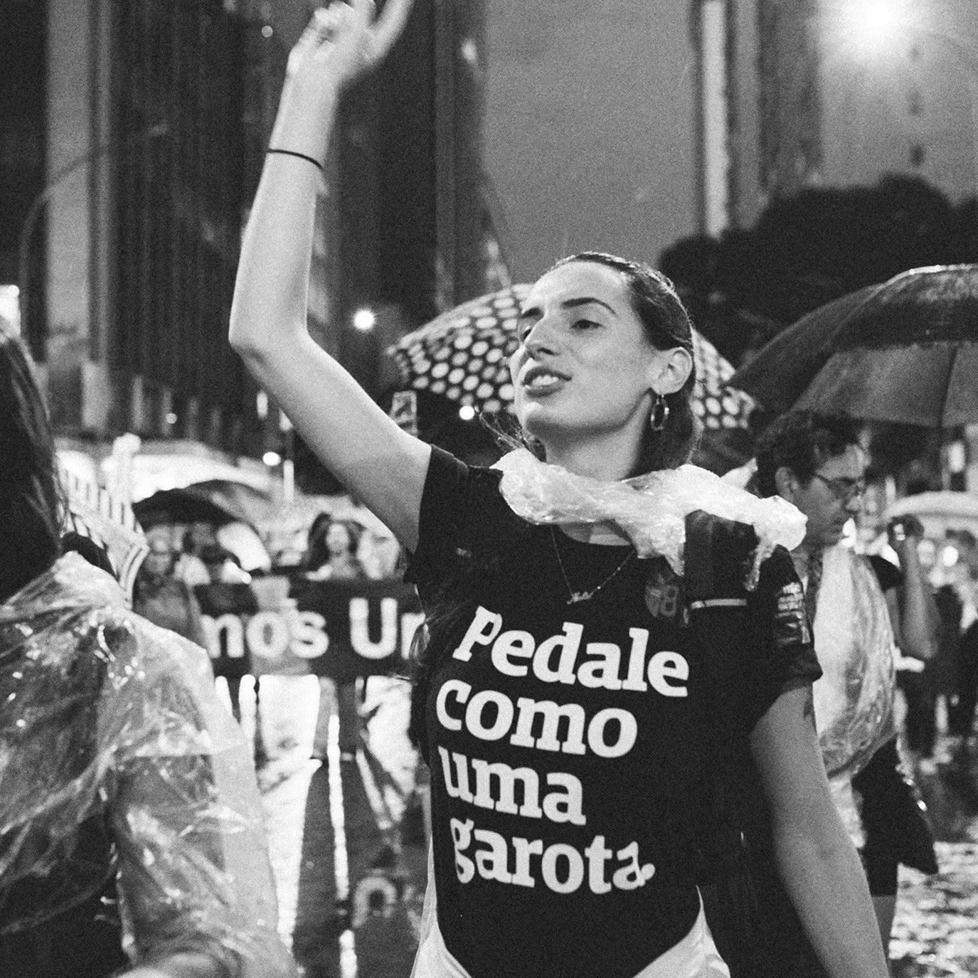 Foto preto e branco de mulher branca de cabelos escuros, veste uma camiseta preta com a frase Pedale como uma garota em branco , com mochila nas costas, está com o braço esquerdo erguido. Está em uma manifestação.