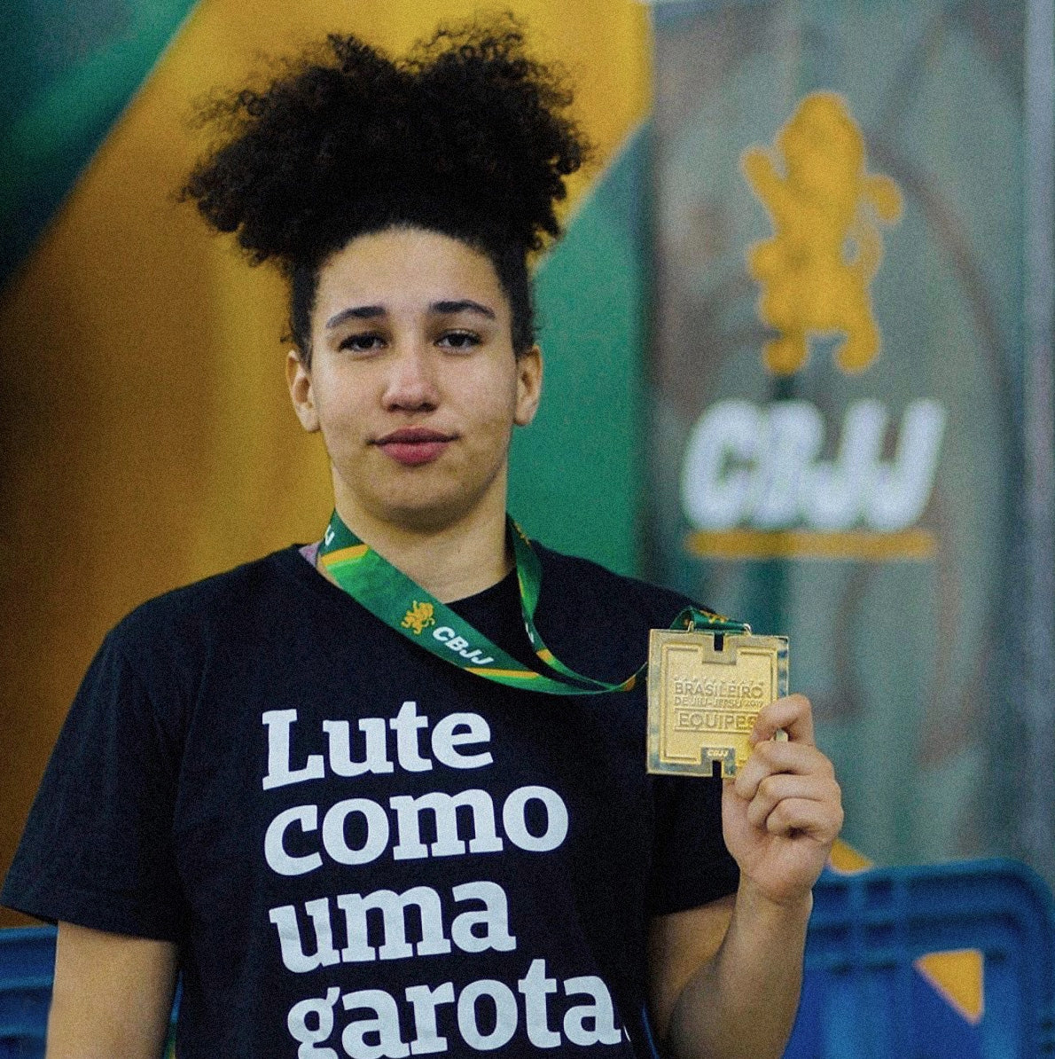 Foto colorida de Maria, mulher negra de cabelos crespo presos, medalhista em jiu jitsu, veste camiseta preta com frase Lute como uma garota em branco,  segura com a mão direita uma medalha com cordão no pescoço. Fundo desfocado