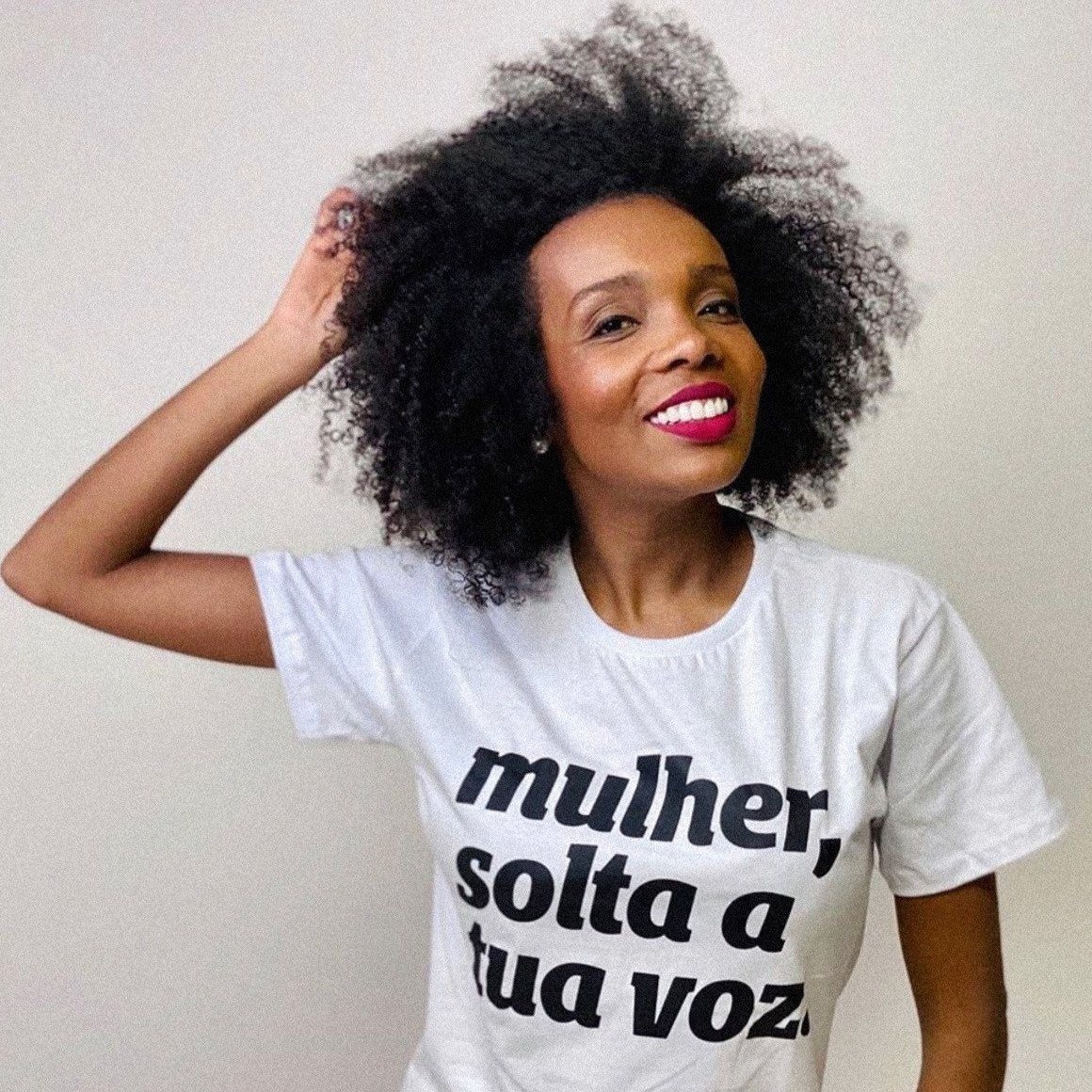 Foto colorida da Thelminha, uma mulher negra, cabelos castanhos, crespos, médios, soltos, usa camiseta branca escrita mulher, solta a sua voz em preto. Ela está com a mão esquerda pegando nos cabelos.