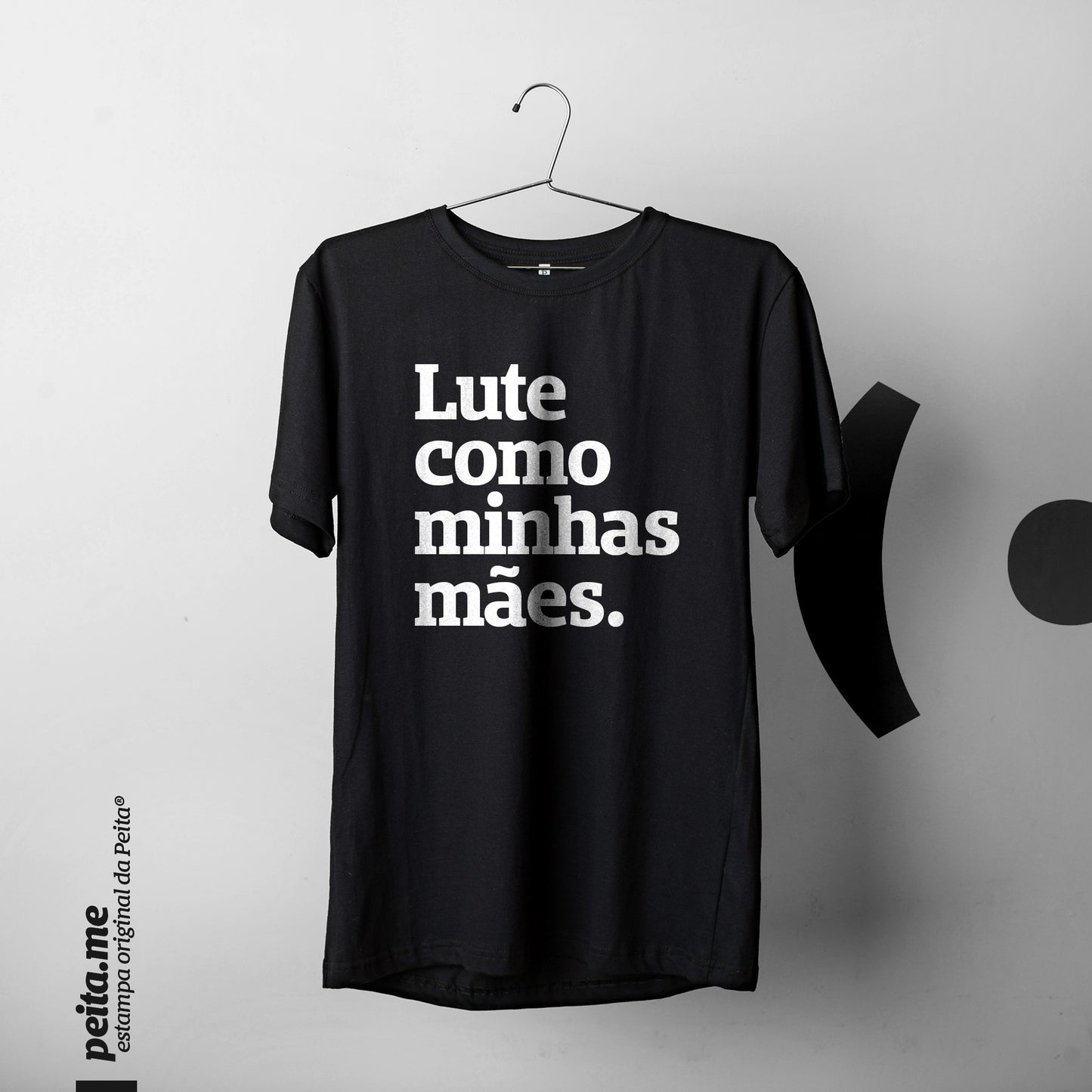 Foto colorida de uma camiseta preta com a frase Lute como minhas mães em branco, pendurada no cabide. Fundo cinza