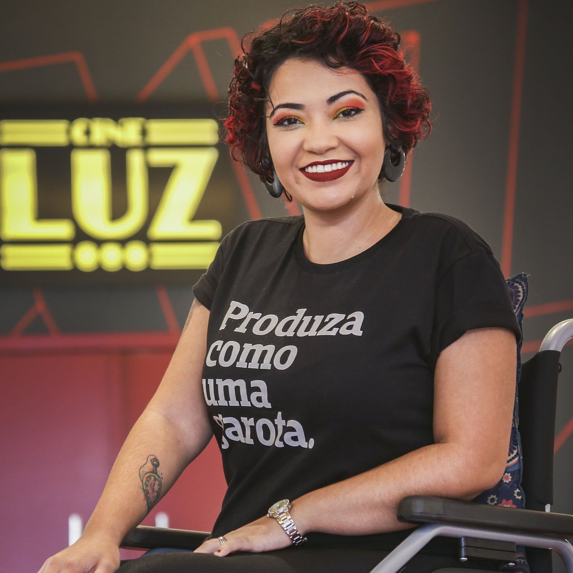 Foto colorida de uma mulher branca, cabelos castanhos, cacheados, curtos, com mechas vermelhas, relógio no pulso esquerdo, tatuagem no braço direito, está vestida com camiseta preta escrita produza como uma garota em branco. Ela é cadeirante e sorri na foto.