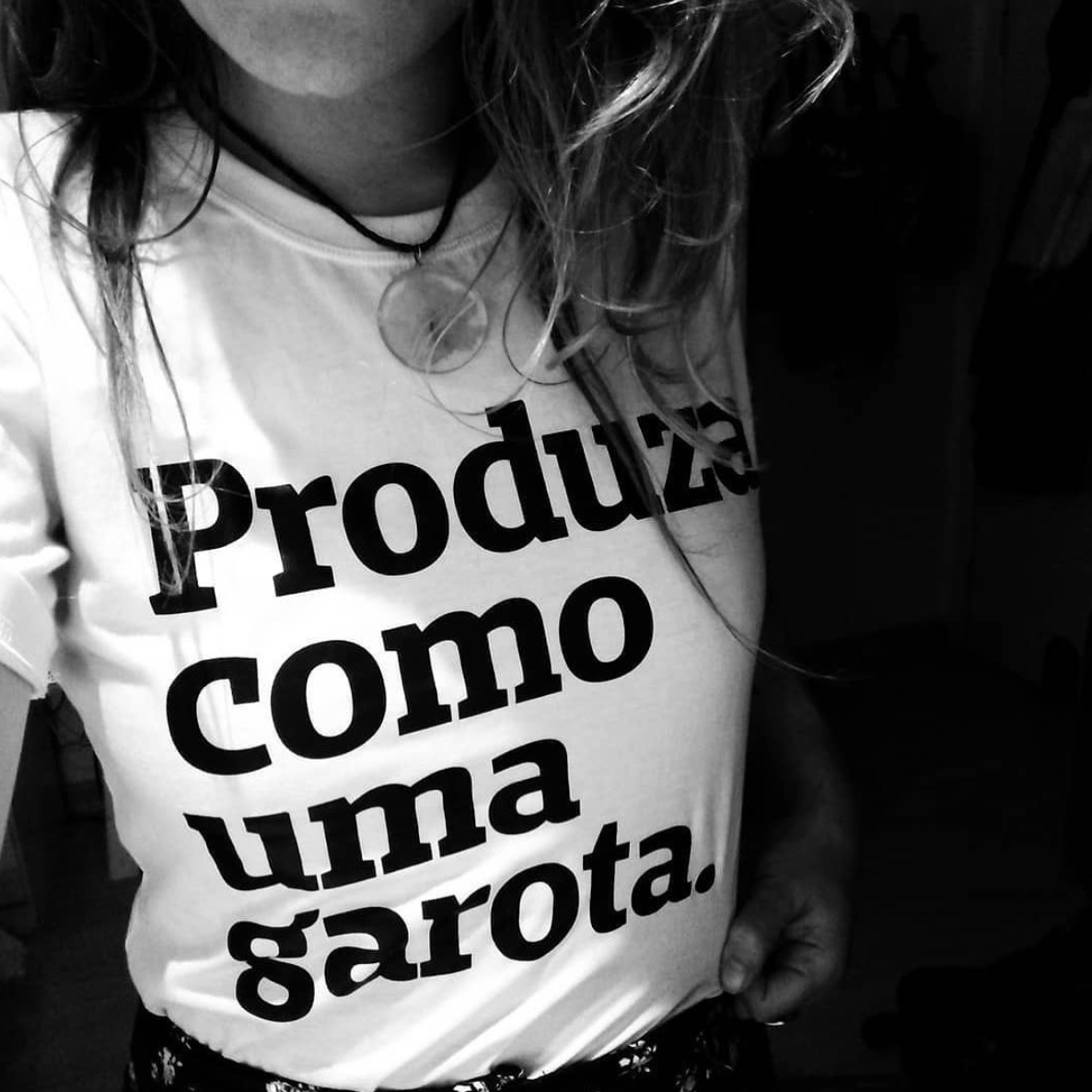 Foto selfie tronco de uma mulher branca, cabelos castanhos claros, lisos, soltos, com colar no pescoço,vestida com camiseta branca escrita produza como uma garota em preto. Ela puxa a camiseta pra baixo com a mão direita, não aparece o rosto.