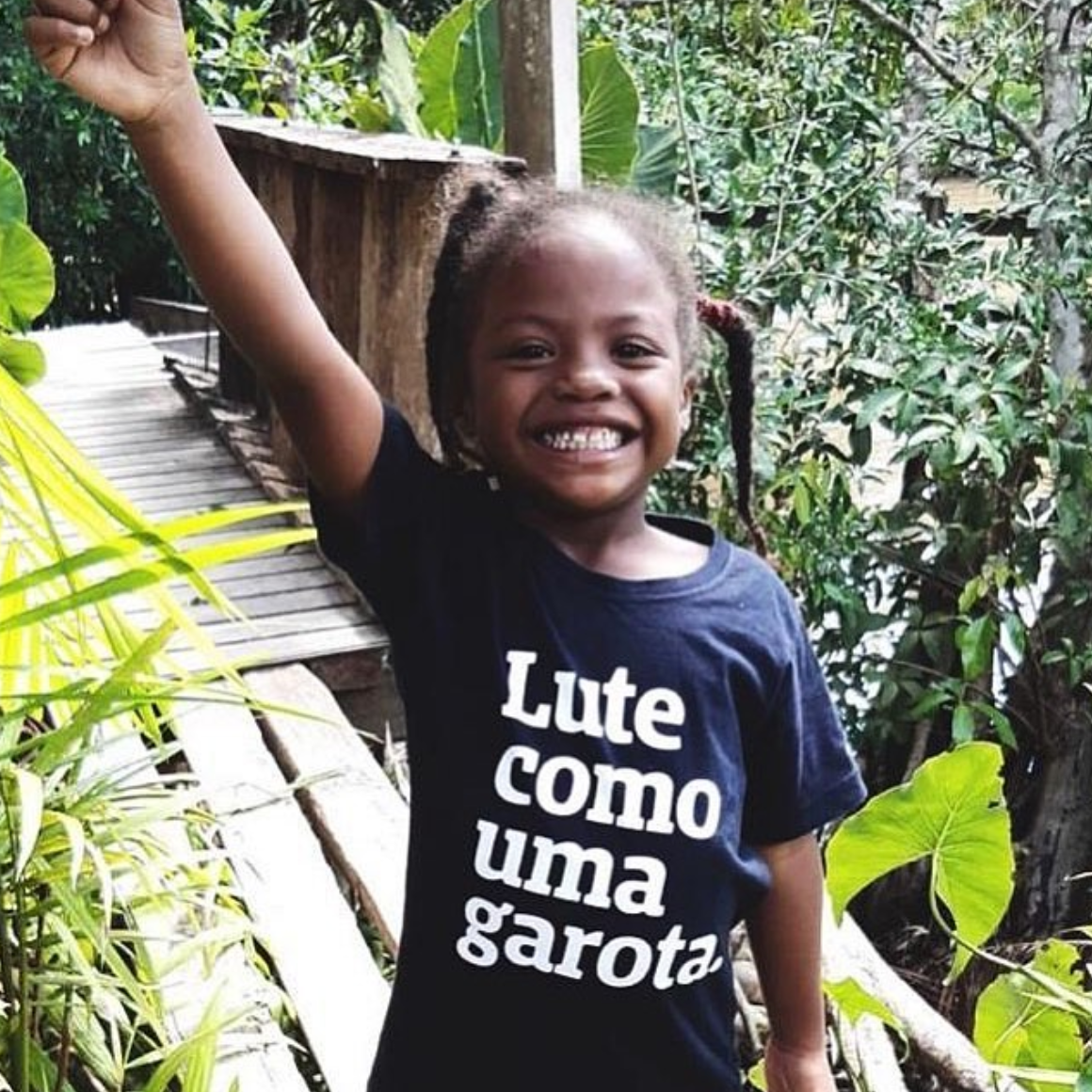 Foto colorida de uma menina negra, cabelos castanhos, com 2 trancinhas, está vestida com camiseta preta escrita lute como uma garota em branco. Está com punho esquerdo fechado e levantado.