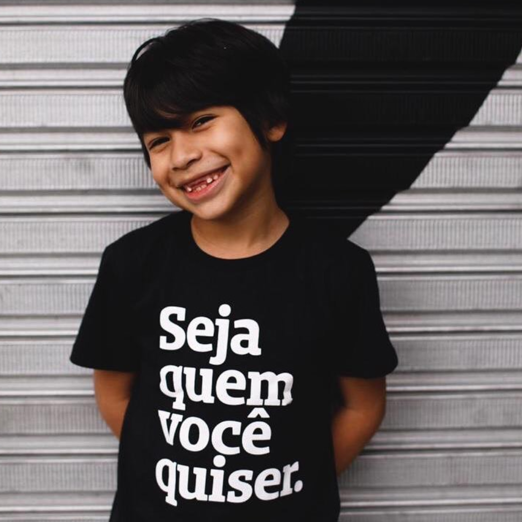 Foto colorida de um menino indígena, cabelos castanhos, lisos, curtos, veste camiseta preta escrita seja quem você quiser em branco. Ele sorri na foto.