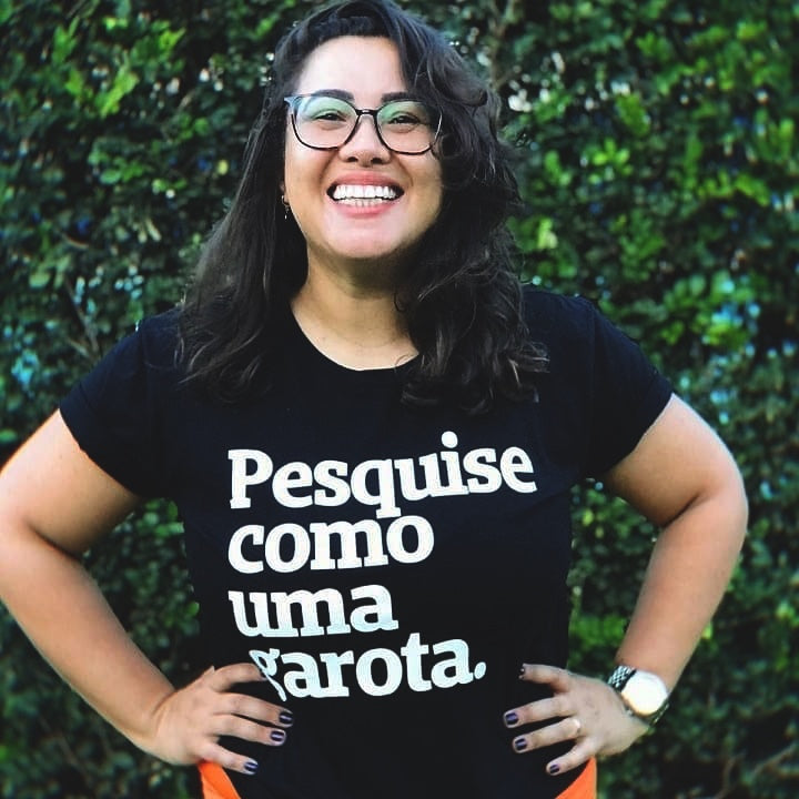 Foto colorida de uma mulher branca, cabelos castanhos, ondulados, médios, soltos, usa relógio no pulso direito e está vestida com camiseta preta escrito pesquise como uma garota em branco. Ela está com as mãos na cintura e sorrindo para foto.