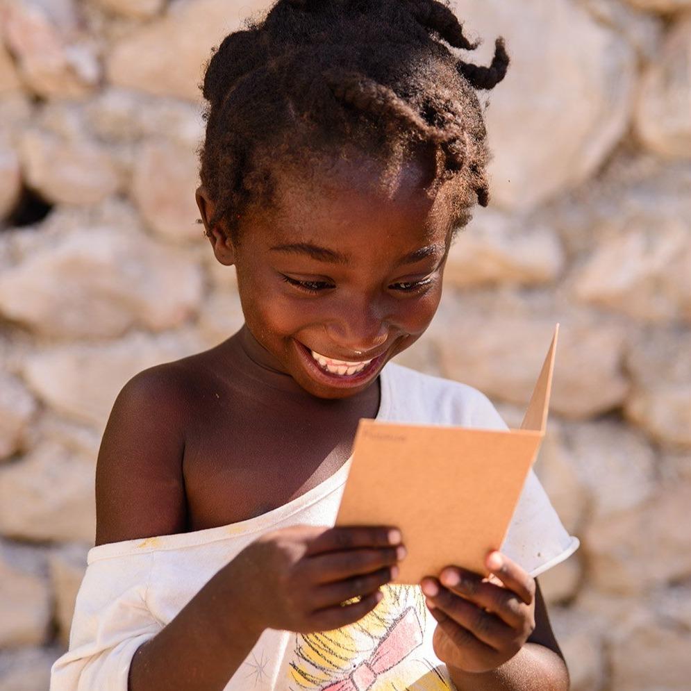 Foto colorida de uma criança negra, cabelos crespos, castanhos, com trancinhas. Ela olha para um cartão nas mãos e sorri