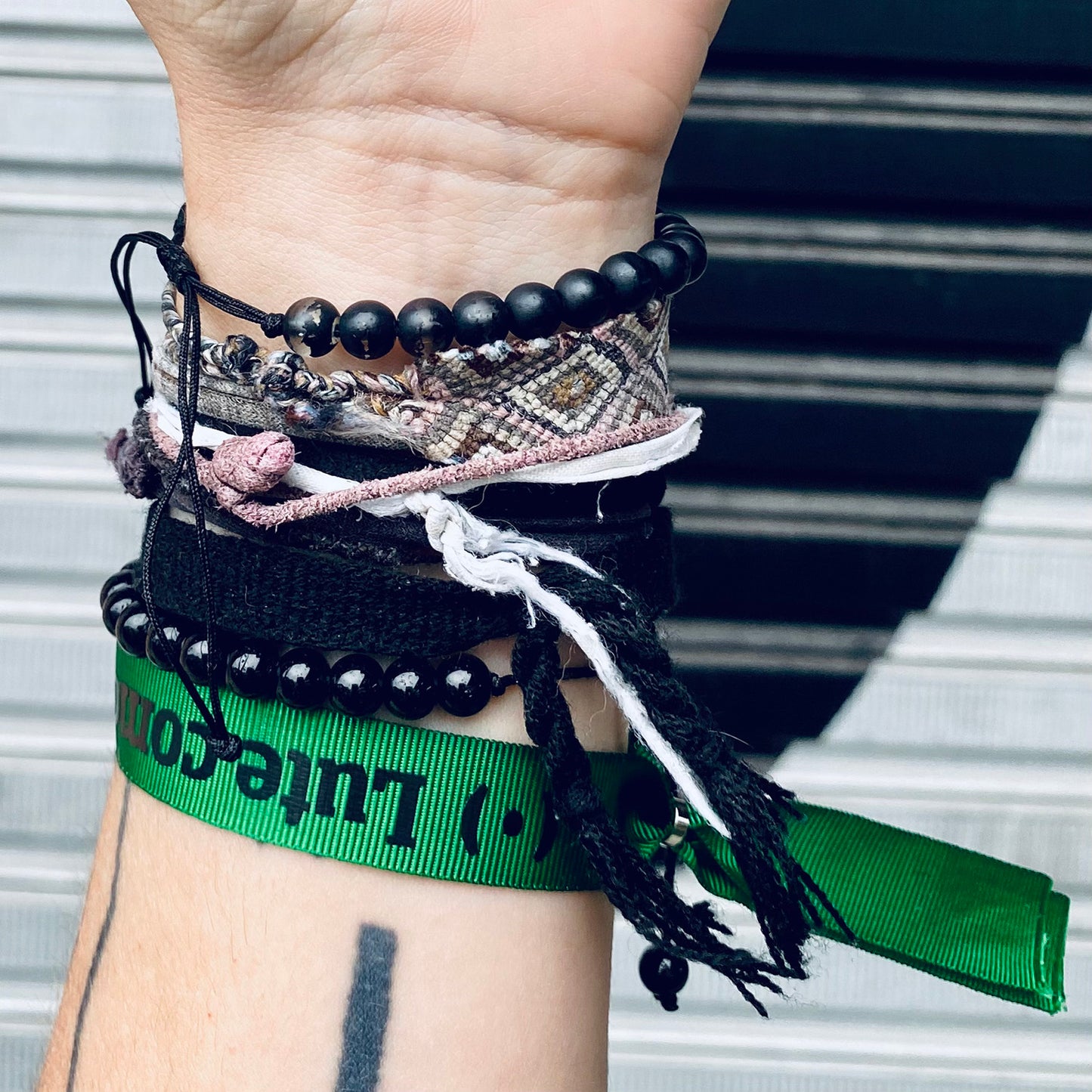 Foto colorida de um braço de pele branca, com uma pulseira verde escrito Lute como em preto e outras pulseiras de bolinhas e macramê. Fundo porta de ferro.